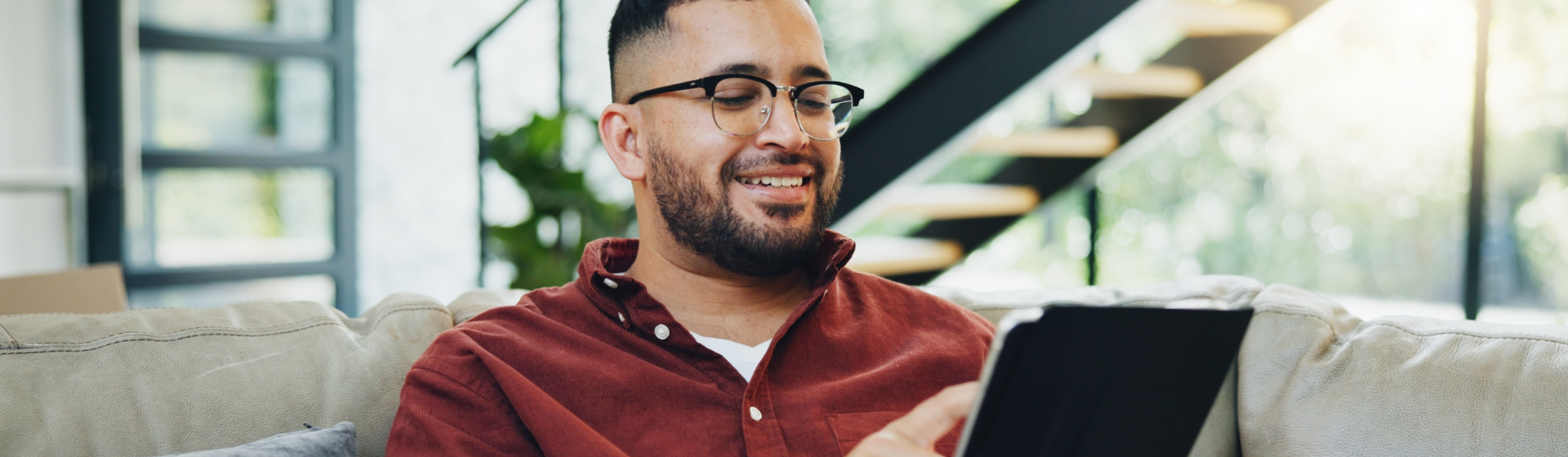 Hispanic male with reading glasses , reading off of a digital tablet with sun coming in through glass window