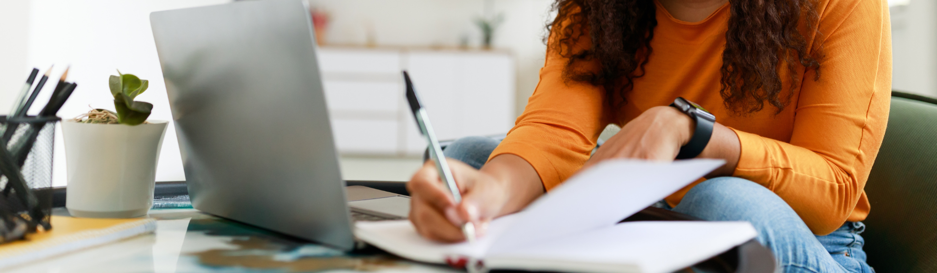 Young black woman with curly hair sitting at desk working on laptop writing notes in paper notebook