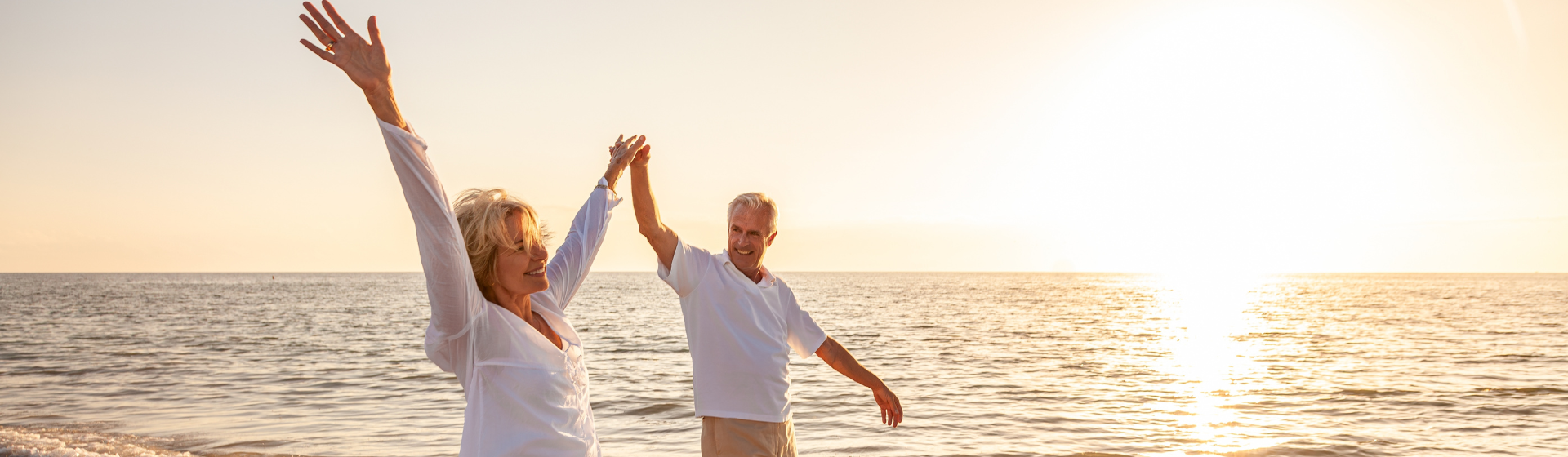 Happy senior man and woman old retired couple walking and holding hands arms raised on a beach at sunset.