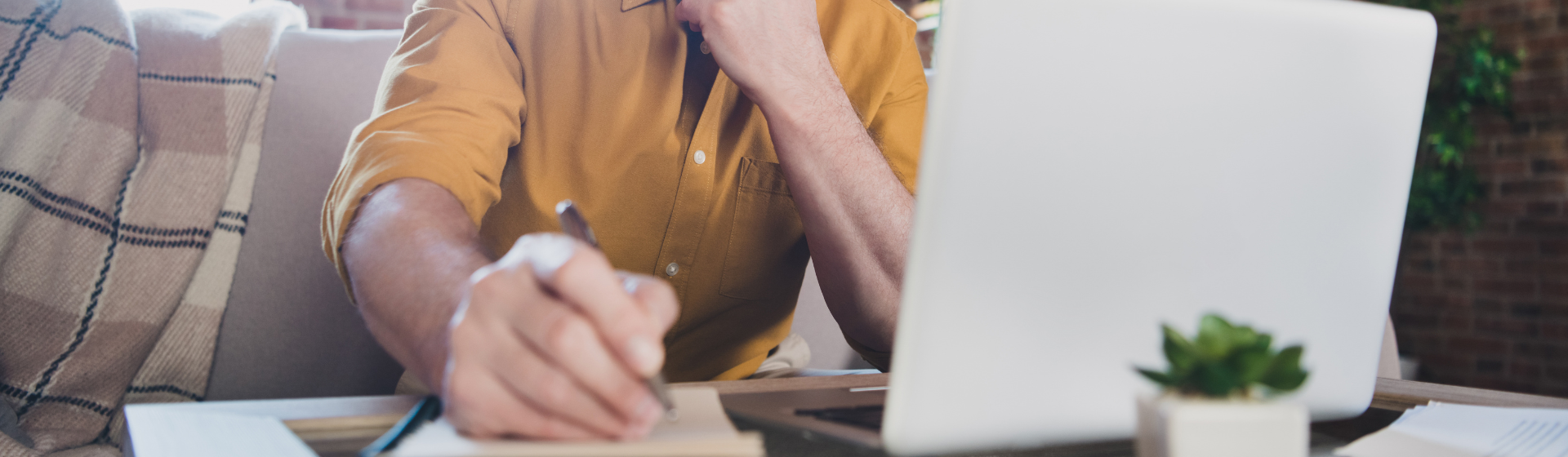 Older caucasian man writing down notes while looking at computer on couch