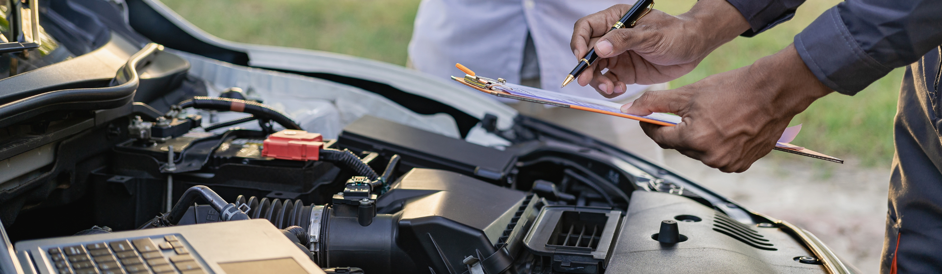 Mechanic with clipboard and computer inspecting under the hood of a vehicle
