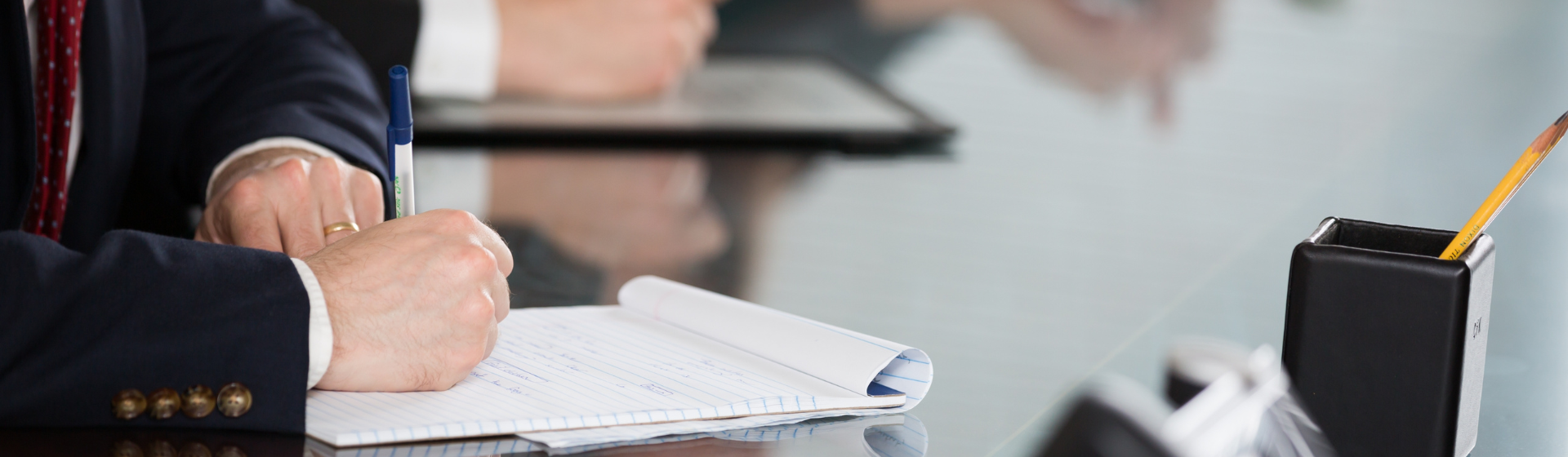 Caucasian man with business attire writing down notes in a professional meeting ; blurred background shows other individuals in meeting